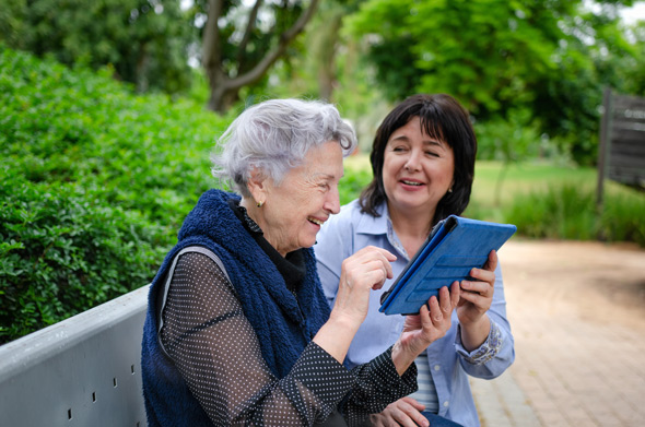 Two women sitting on a bench outside, engaged in a joyful conversation while looking at a tablet. The scene is set in a lush, green park, highlighting their friendly interaction and shared moments of fun.