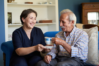 A cheerful woman hands a cup of tea to an elderly man while sitting together in a cozy living room, showcasing a warm and caring interaction.