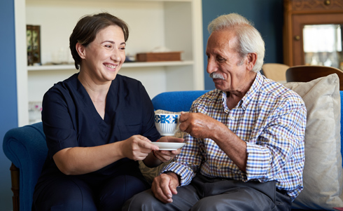 A caregiver and an elderly man sharing a moment, both smiling while enjoying tea in a comfortable home setting.