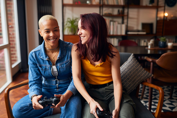 Two friends sitting on a couch, smiling and holding video game controllers, in a cozy living room setting.