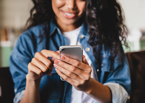 A woman with curly hair wearing a denim jacket, smiling while using her smartphone.