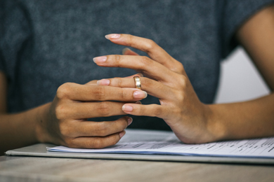 close-up of a person's hands resting on a table, wearing a gold ring, with neatly manicured nails, and a stack of papers beneath them