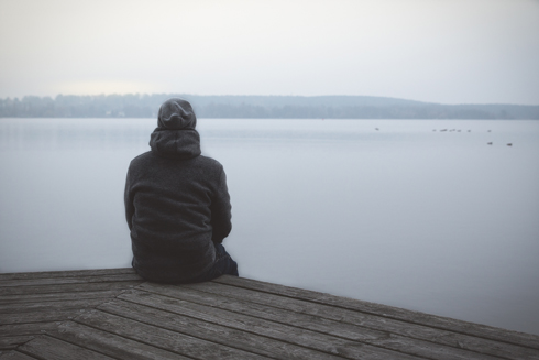 A person sitting on a wooden dock by a tranquil lake, gazing at the calm water and distant hills under a gray sky.