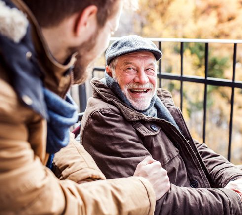 A smiling elderly man in a brown coat and gray hat enjoys a joyful conversation with a younger man, both seated outdoors with a backdrop of autumn foliage.