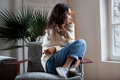 Woman sitting on a chair, legs pulled to her chest, looking pensively out a window, with indoor plants in the background. Cozy lifestyle moment.