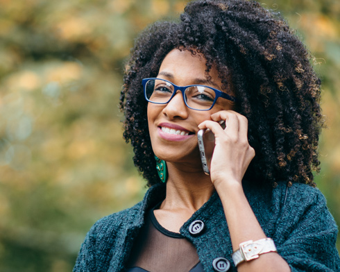 A woman with curly hair and glasses smiles while talking on a smartphone, dressed in a stylish outfit against a blurred outdoor background.