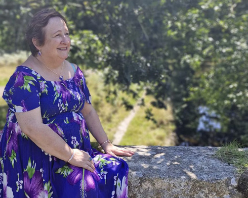 woman in a blue and purple floral dress sitting on a stone wall, smiling under green trees, enjoying a sunny day